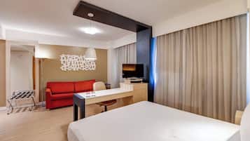A hotel room with a bed, a desk with a TV, a red sofa, and a modern light fixture.