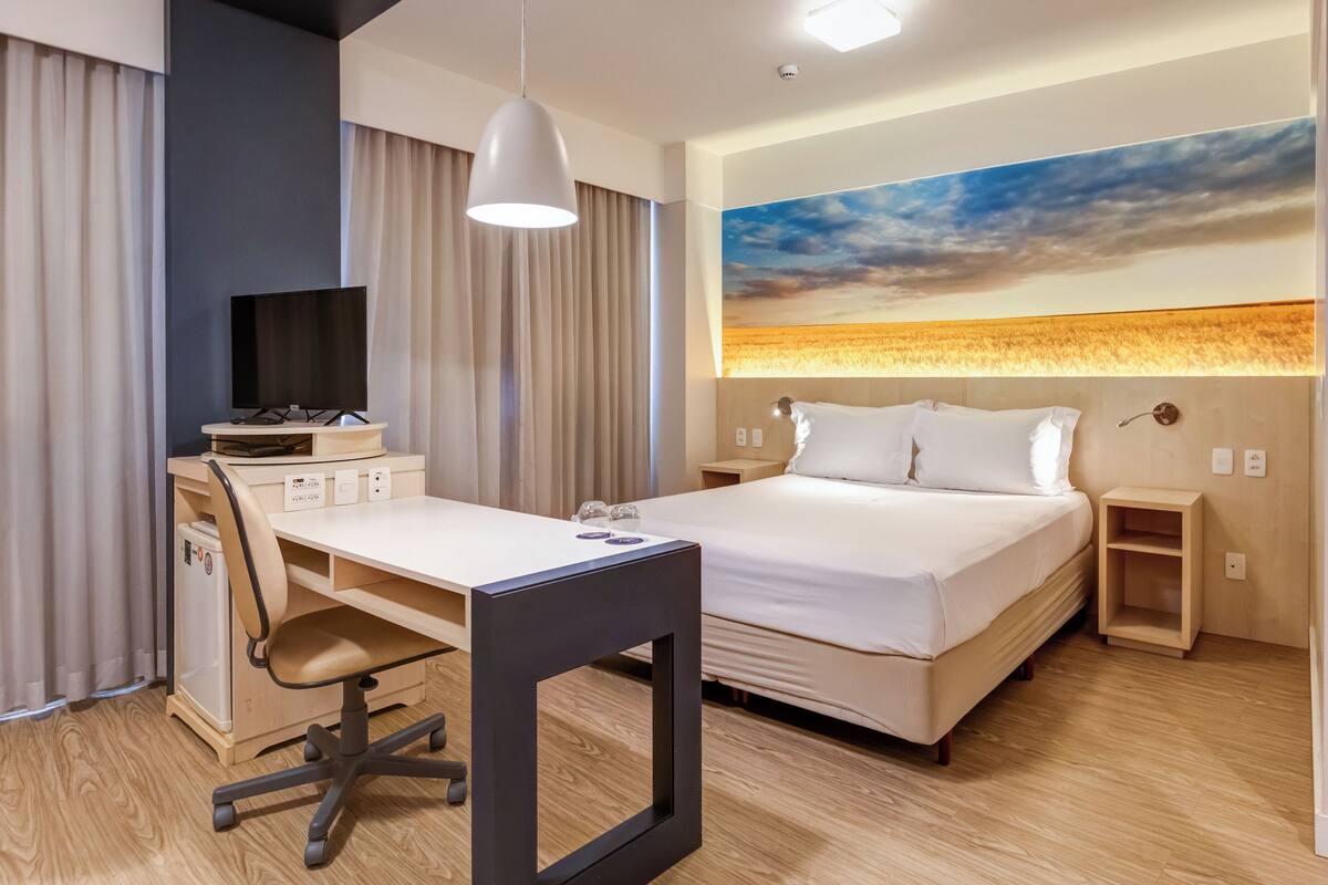 A hotel room with a bed, desk, chair, and a painting of a sunset.