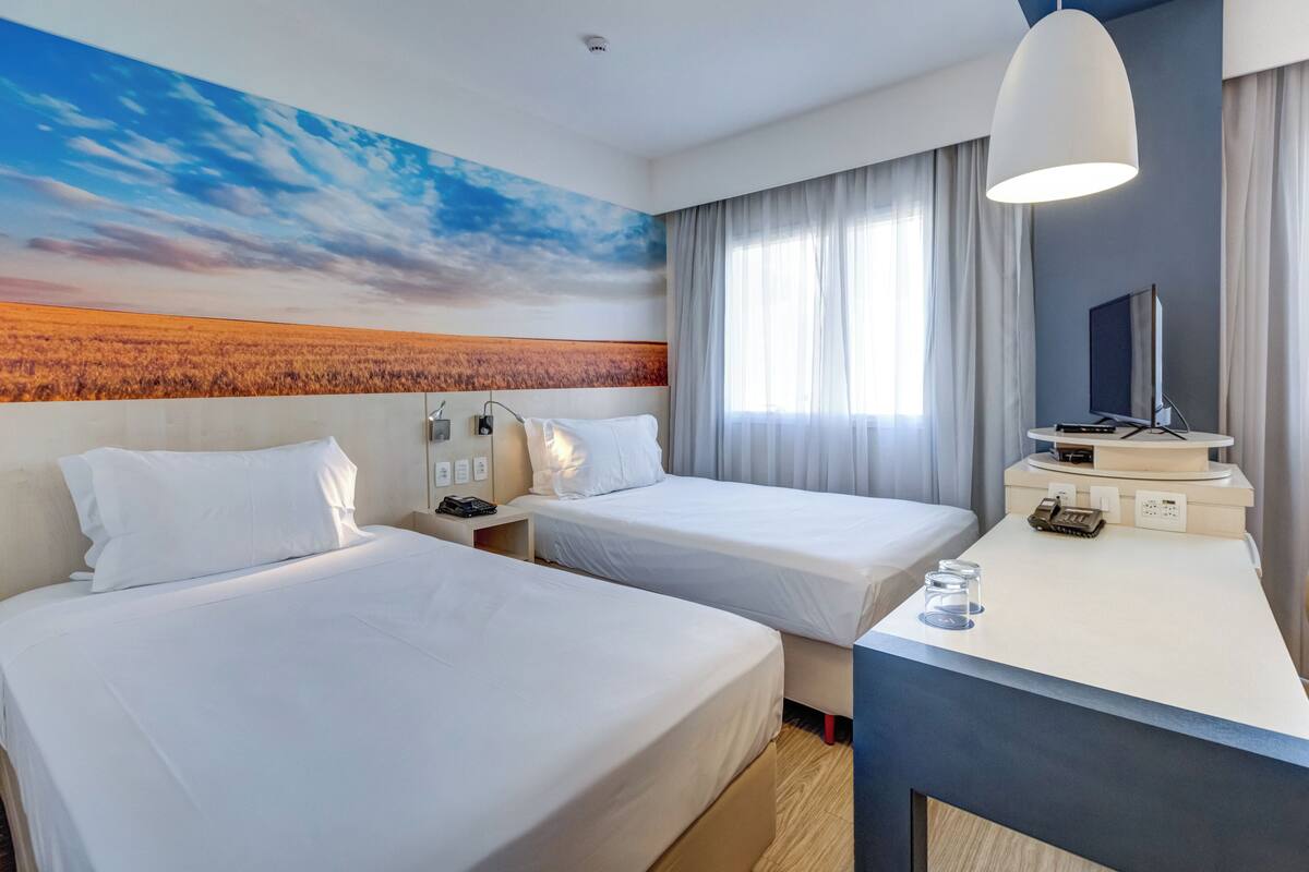 A hotel room with two beds, a desk, a flat-screen TV, and a painting of a sunset over a field.