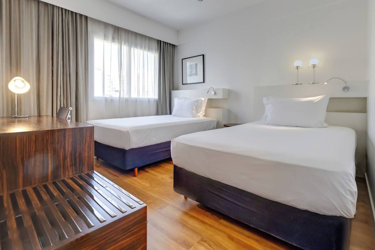 A hotel room with two beds, a wooden bench, a desk, and a large window with curtains.