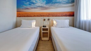 Habitación de hotel con dos camas, una mesita de noche con teléfono y lámpara, y un gran mural en la pared que representa un campo al atardecer.