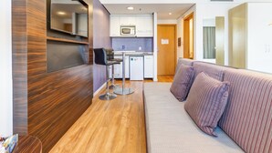 Una habitación de hotel moderna con un revestimiento de pared de madera, un sofá, una zona de cocina con microondas y un pequeño rincón de estar con taburetes.
