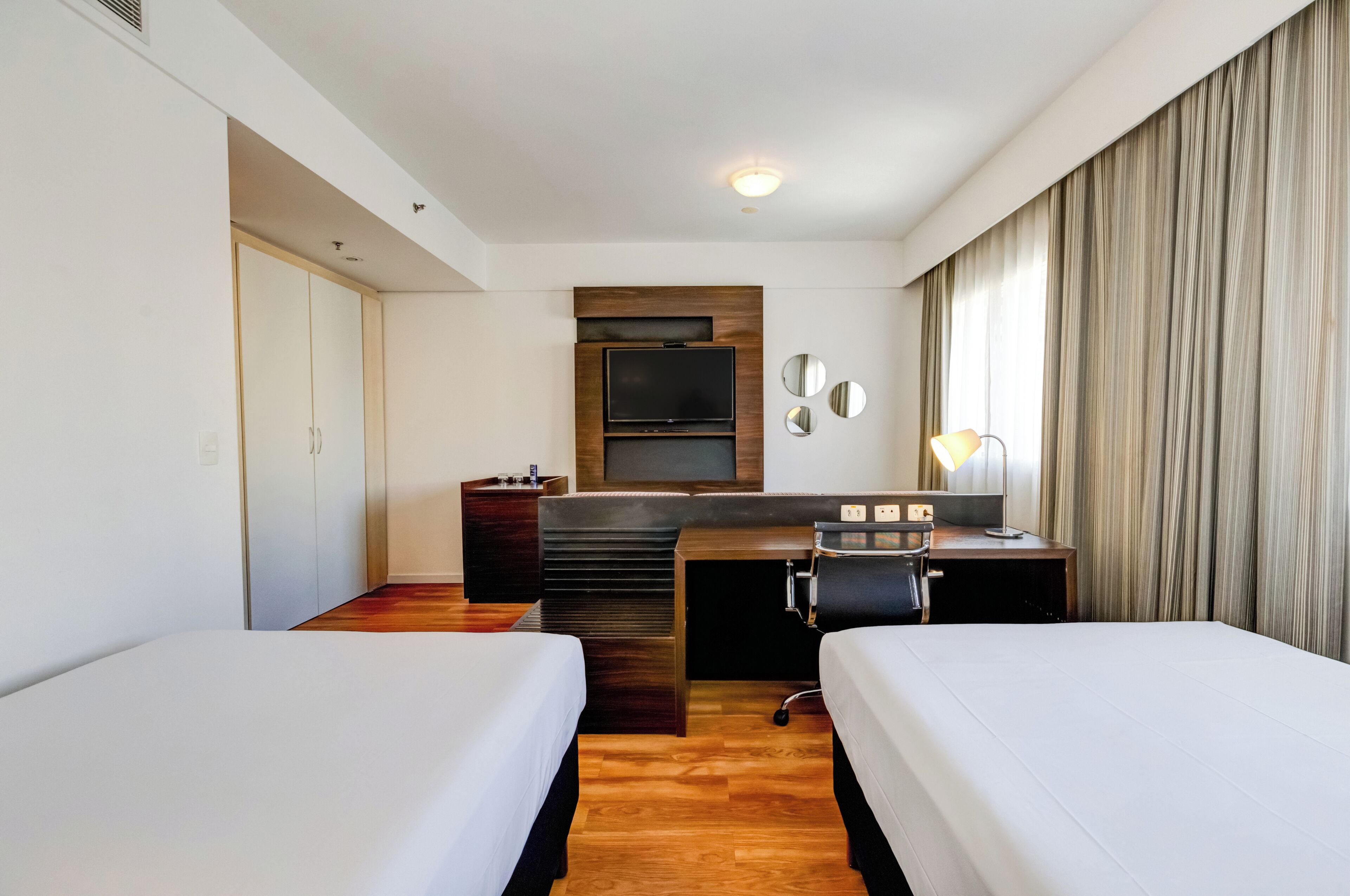 A hotel room with two beds, a desk with a chair, a television, and a large window with curtains.