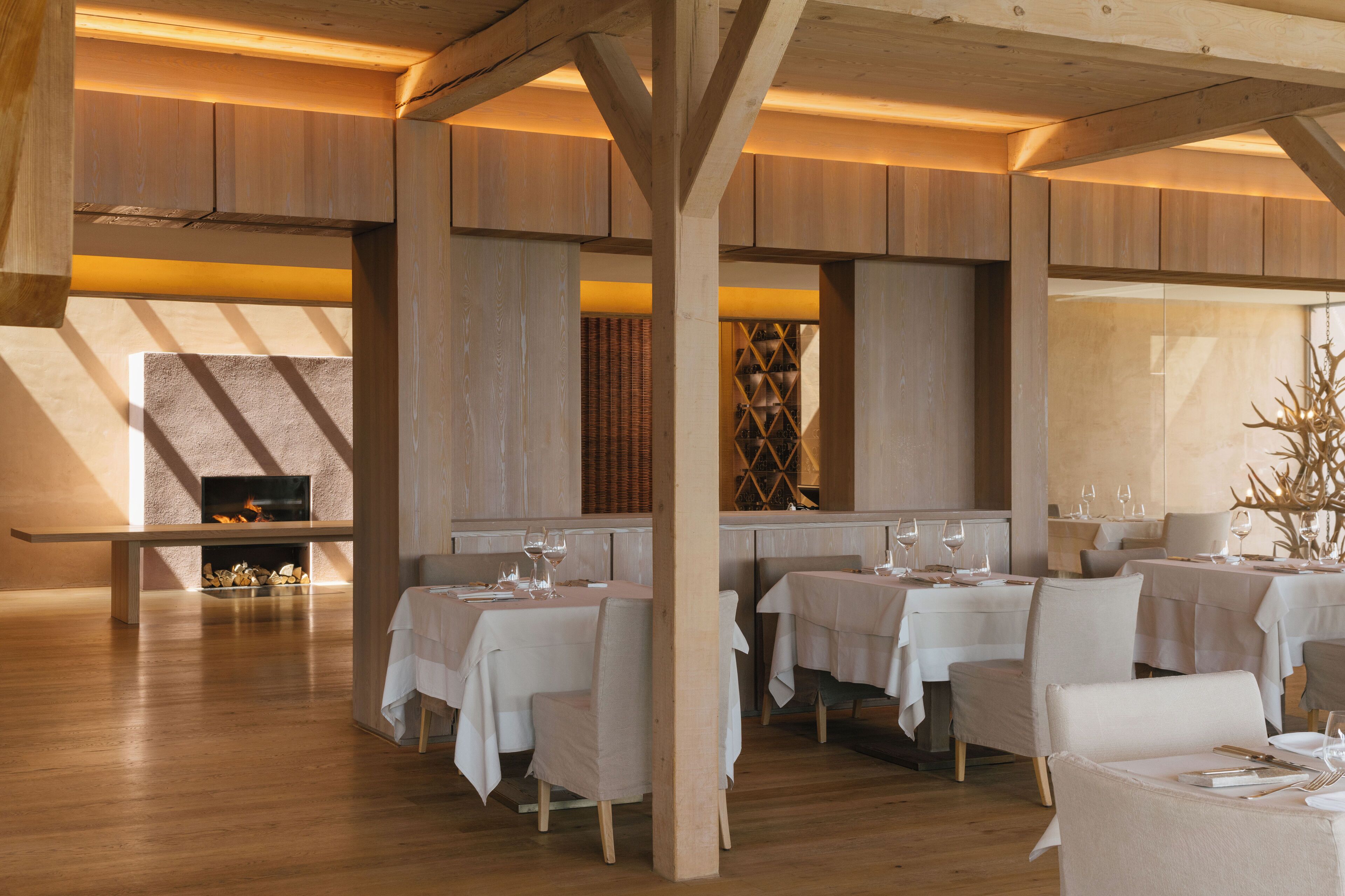 A restaurant with wooden beams, white tables, and chairs. There is a fireplace and a large window.