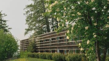 Un edificio moderno con terraza de madera y mesas de picnic en un área ajardinada con árboles y un campo cubierto de césped.