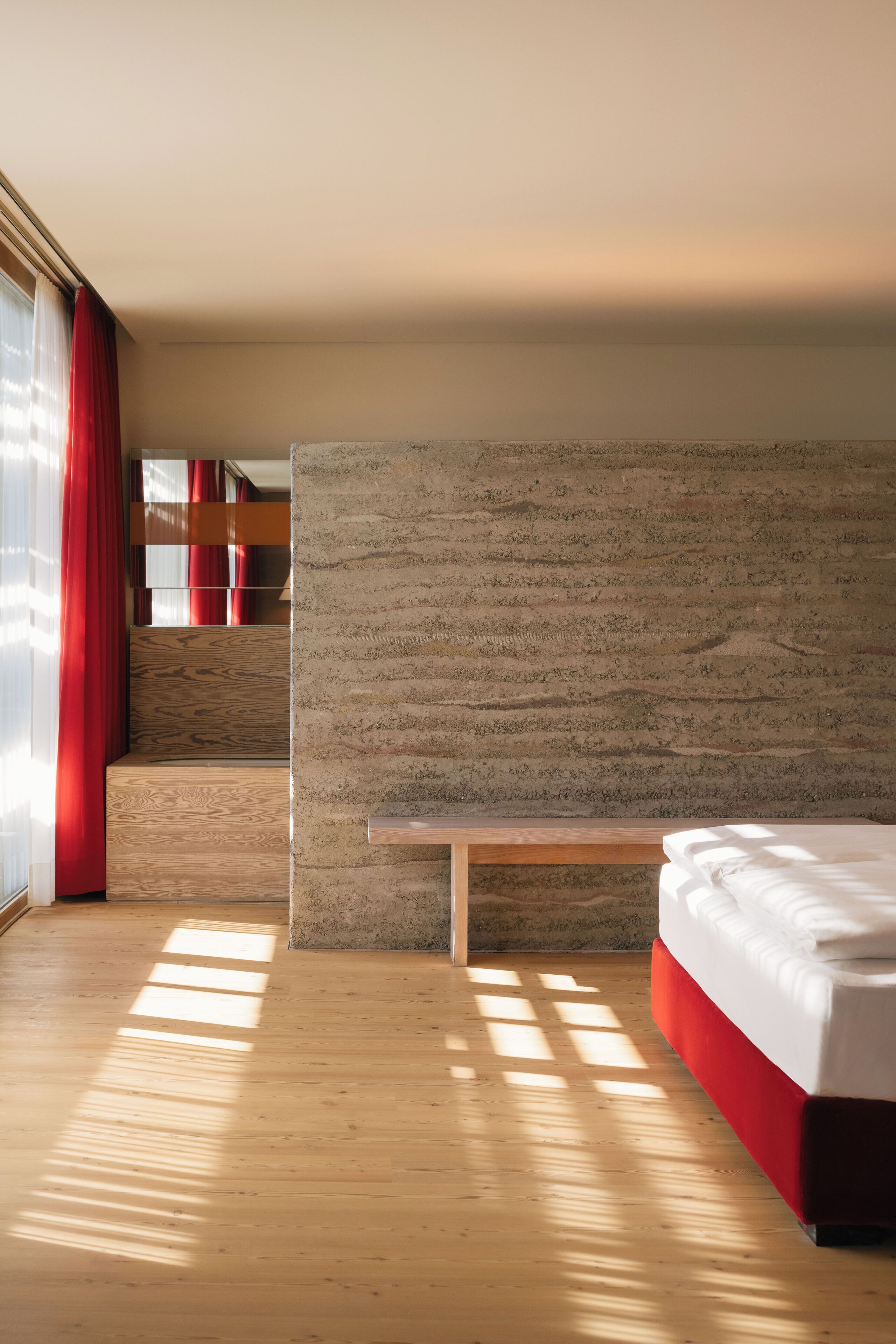 A modern hotel room with a wooden floor, a large bed with a red accent, a stone accent wall, and a view of the city through large windows.