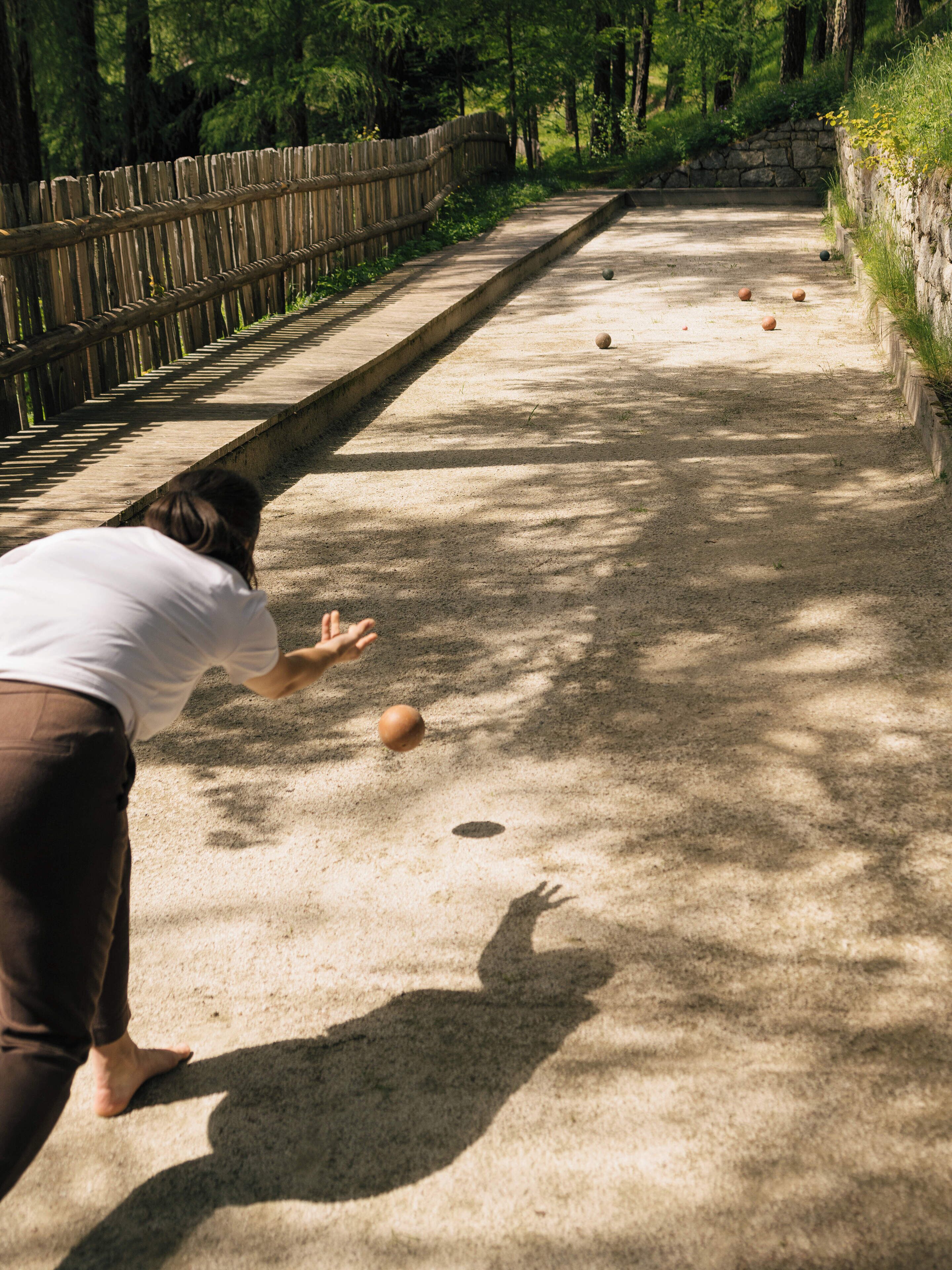 Una persona juega a boliche en un sendero de concreto, con una cerca de madera a un lado y un muro de piedra al otro.
