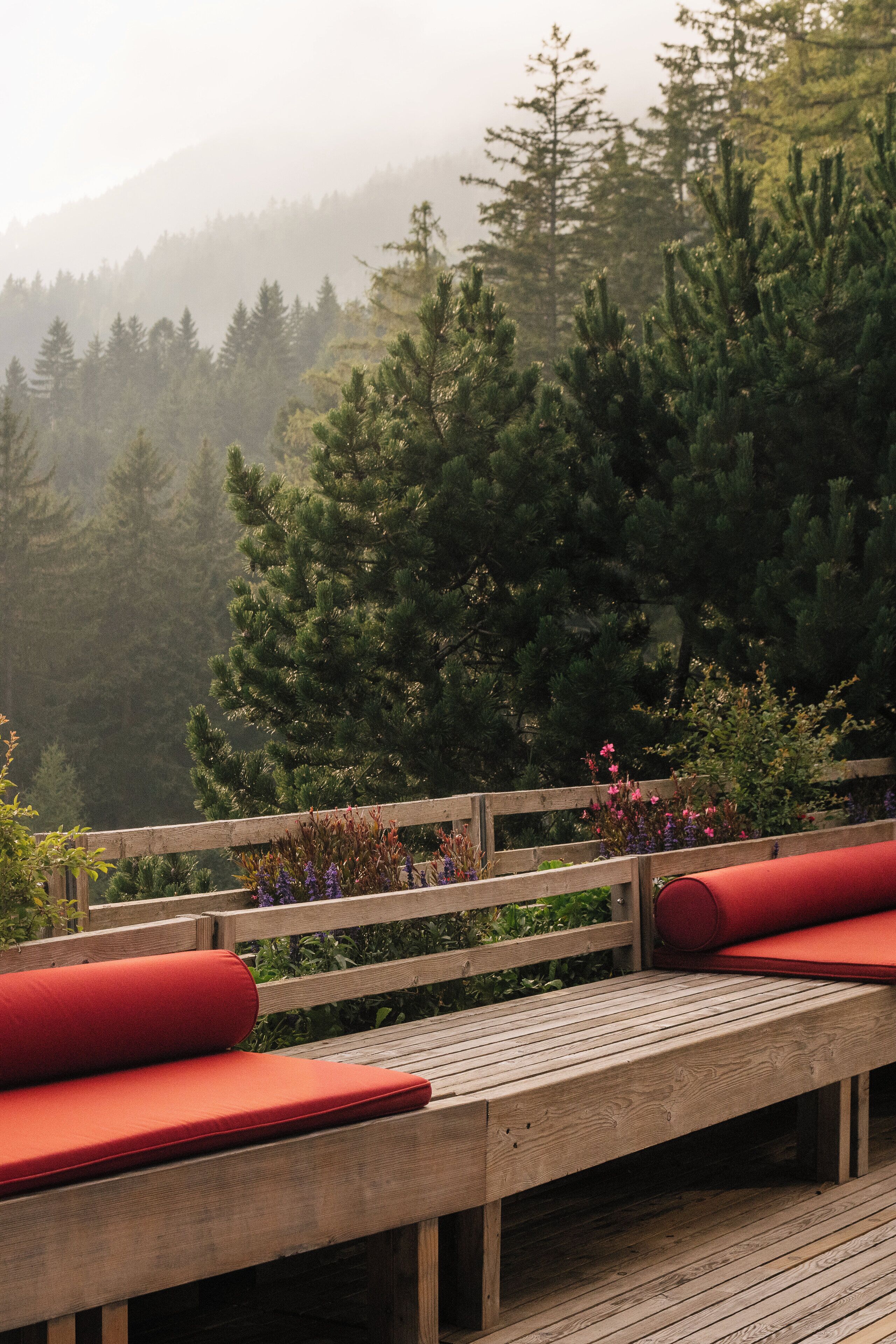 Une terrasse en bois avec des coussins rouges et une balustrade en bois, donnant sur une zone boisée.