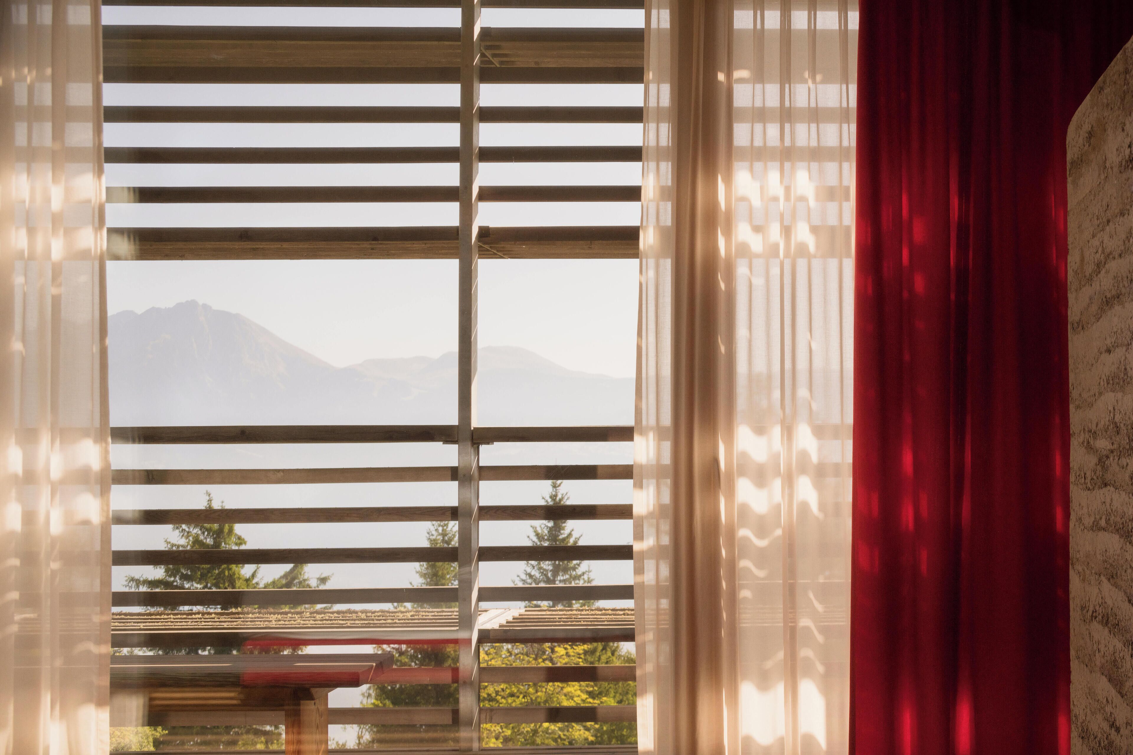 A window with horizontal blinds, a red curtain, and a view of a mountain range.