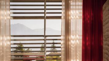A window with horizontal blinds, a red curtain, and a view of a mountain range.