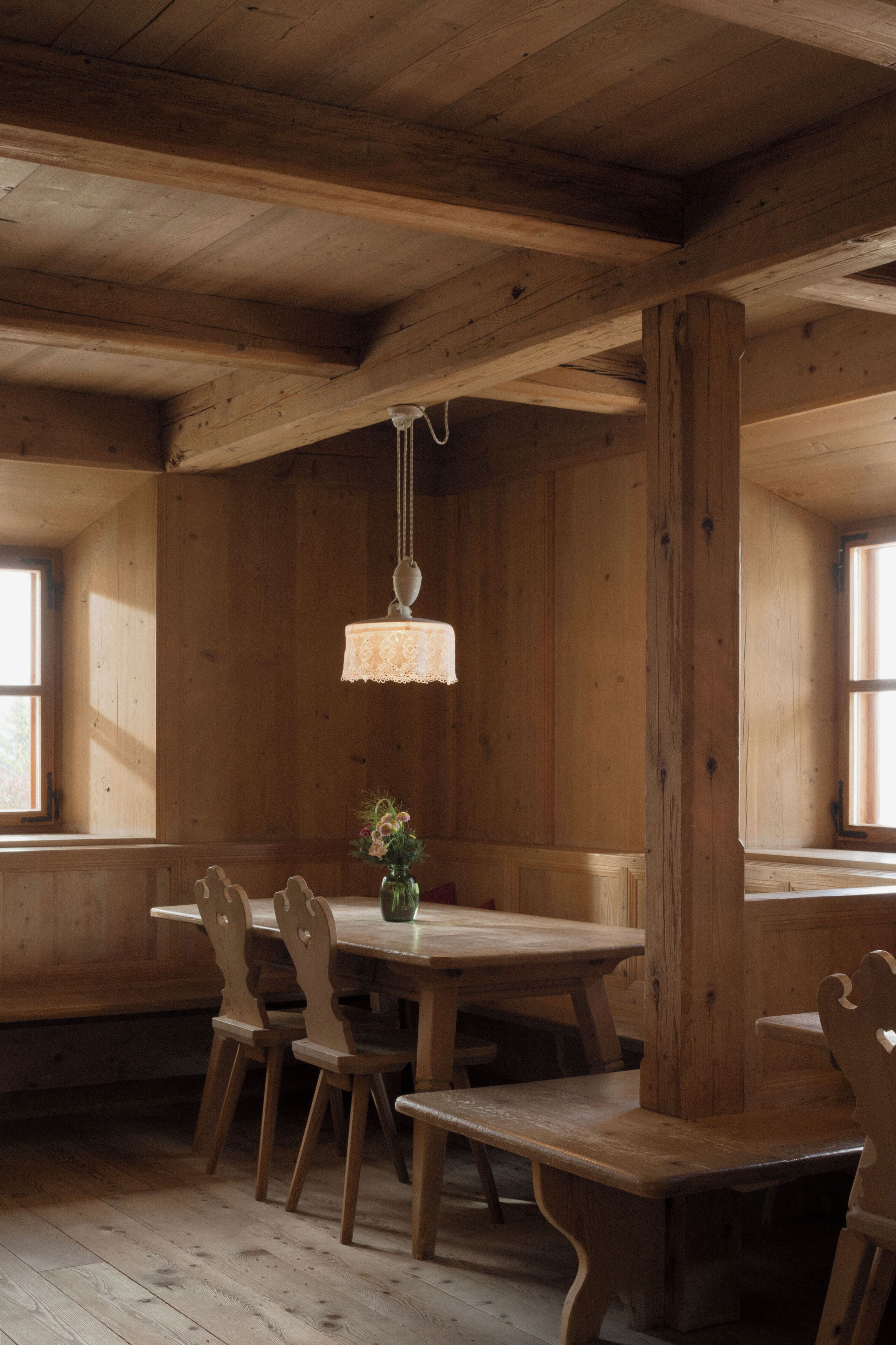 Un comedor de madera con una mesa y sillas, una lámpara colgante y ventanas amplias.