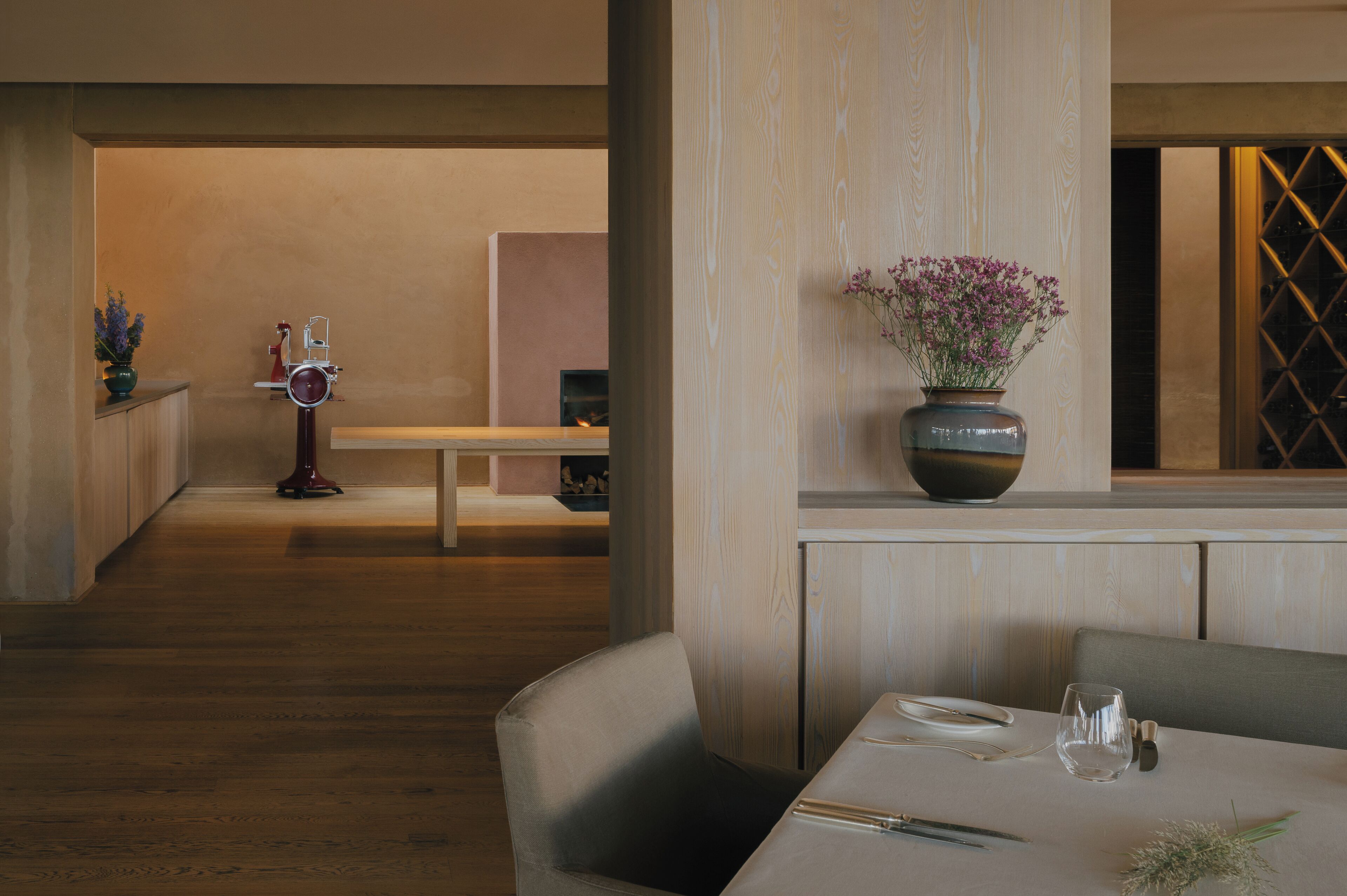 A dining area with a wooden floor, a table set with cutlery, a vase with flowers, and a bench in the background.