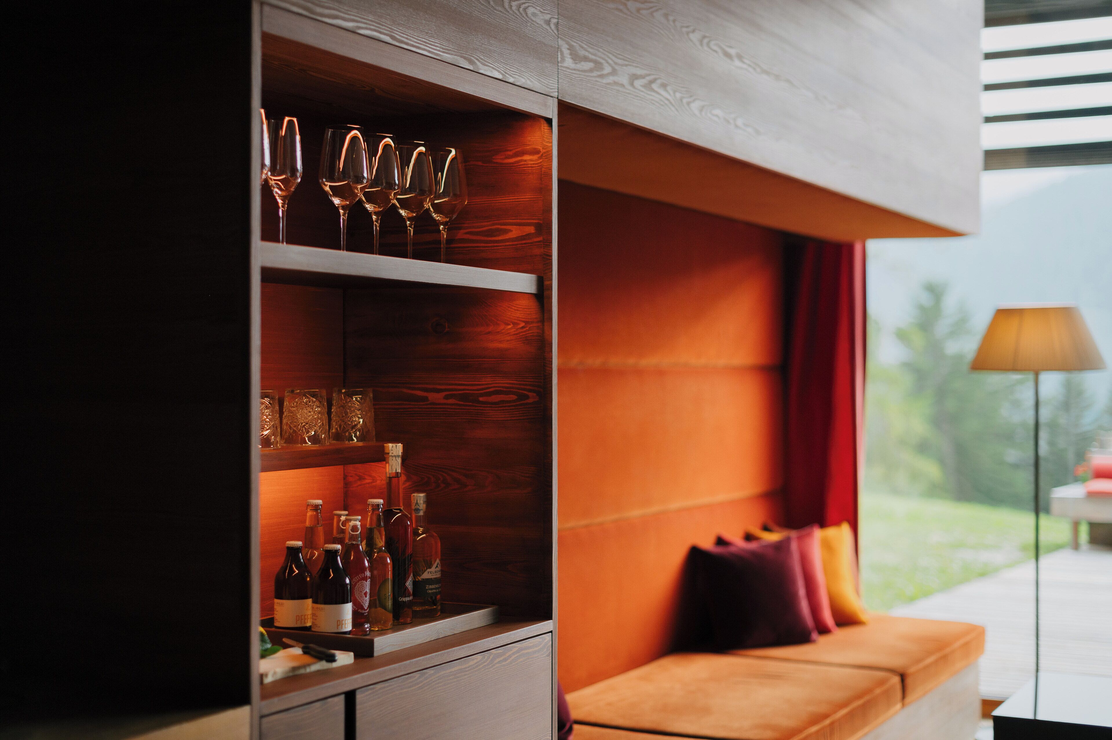 Ein modernes Interieur mit einer Holzkabine, in der Weingläser und Flaschen aufbewahrt werden, einer Couch mit Kissen und Blick auf Grünflächen.