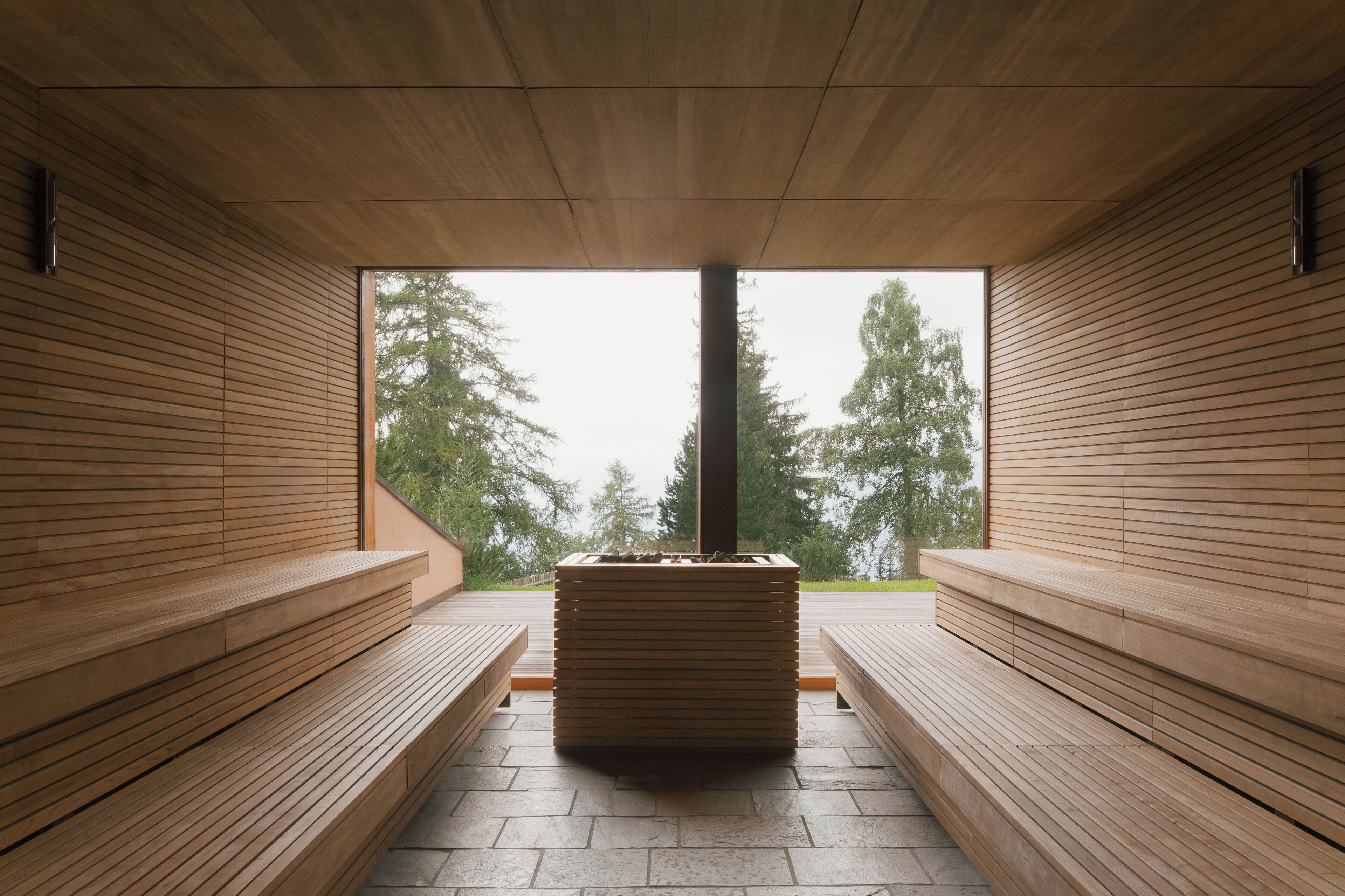 A sauna with wooden benches and a central pillar, large windows with a view of trees.