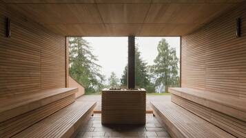 A sauna with wooden benches and a central pillar, large windows with a view of trees.