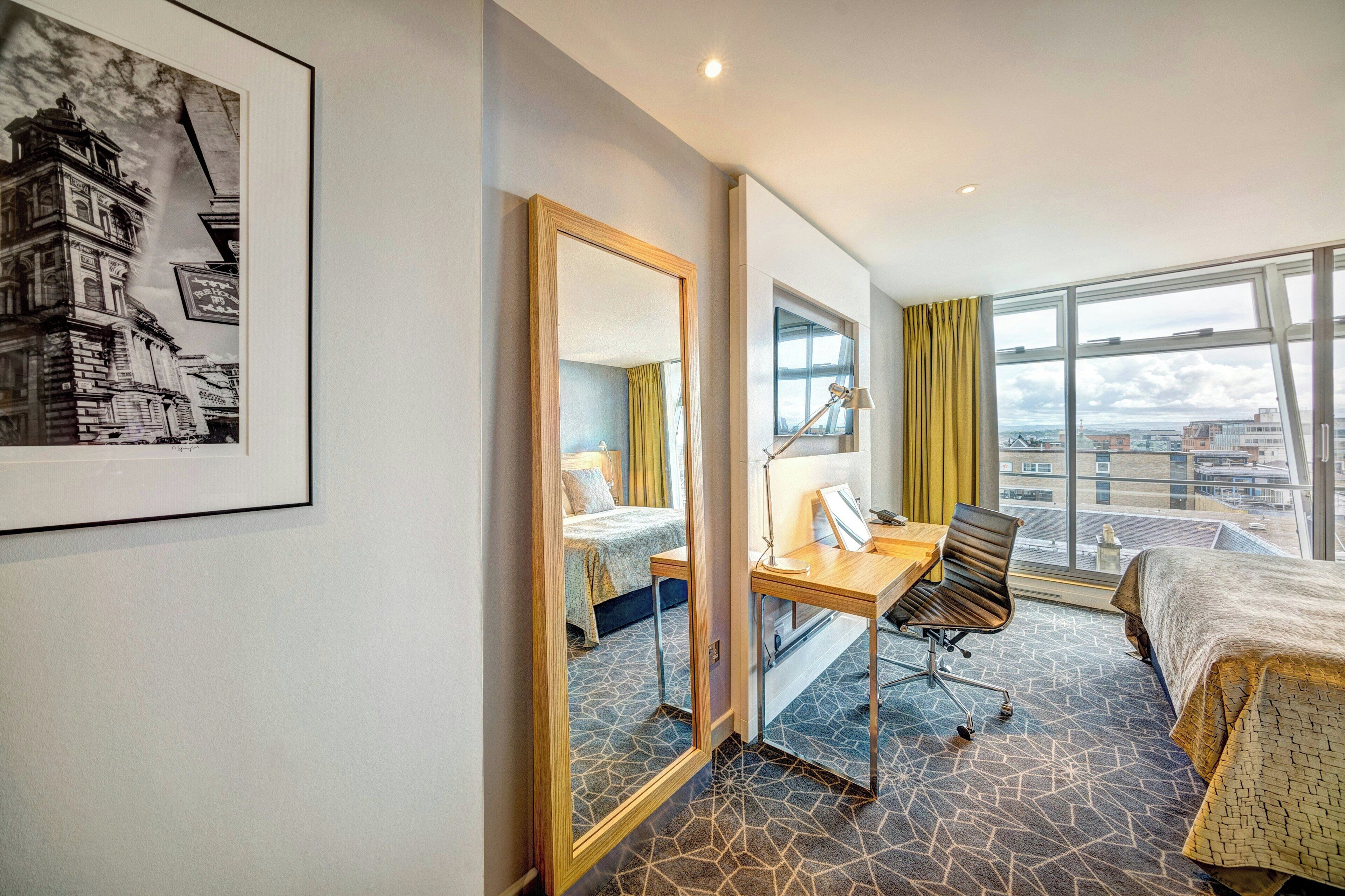 Habitación de hotel con cama, escritorio, silla y ventanas amplias que ofrecen vista a la ciudad.