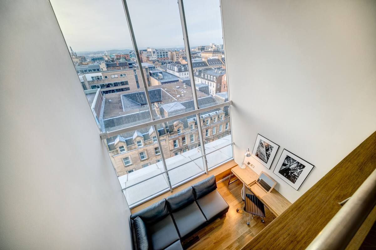 Een hoog gebouwde ruimte met een groot raam met uitzicht op de stad, een zwarte leren bank, een houten bureau en ingelijste foto’s aan de muur.