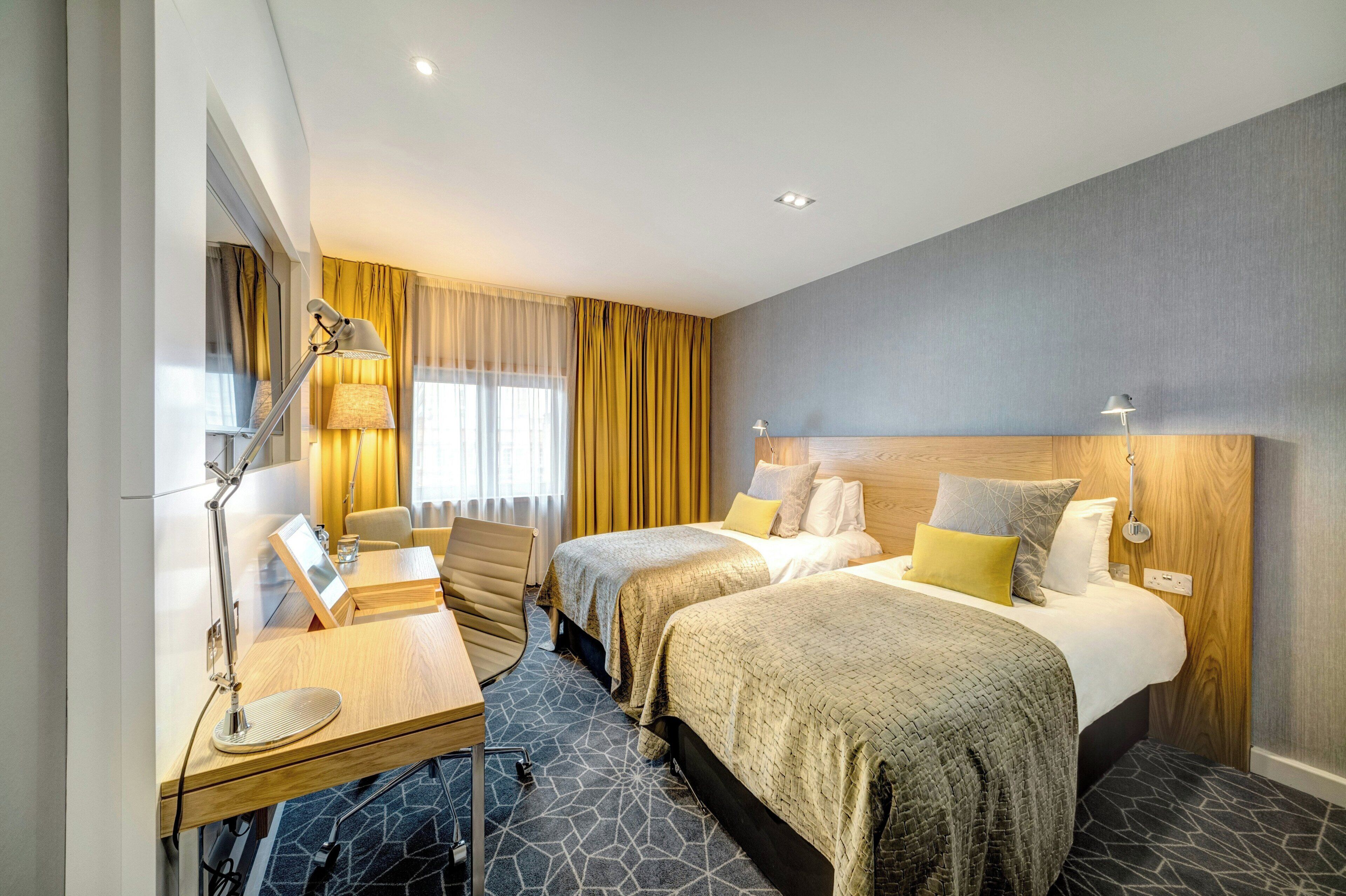 A hotel room with two beds, a desk, a chair, a lamp, and a window with curtains.