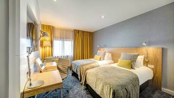 A hotel room with two beds, a desk, a chair, a lamp, and a window with curtains.