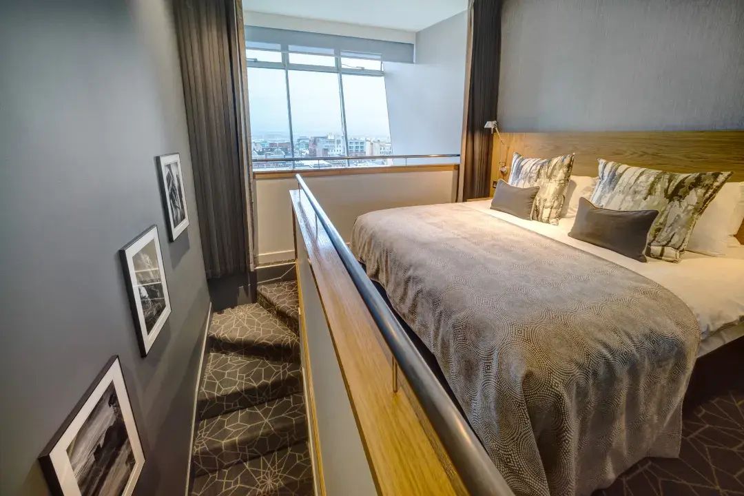 A hotel room with a large bed, a window with a city view, and a staircase with patterned carpet.