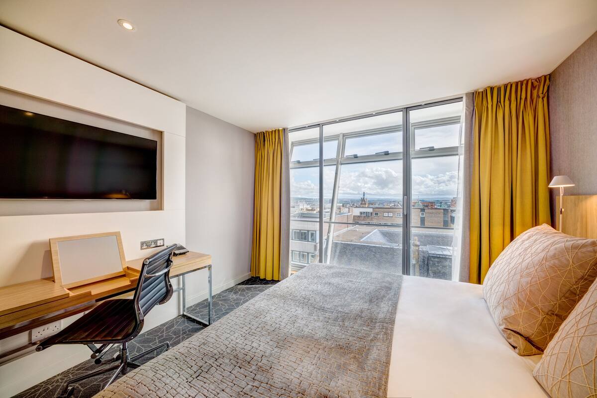 Een moderne hotelkamer met een groot bed, een bureau met een computer, een aan de muur bevestigde televisie en uitzicht op de stad door grote ramen.