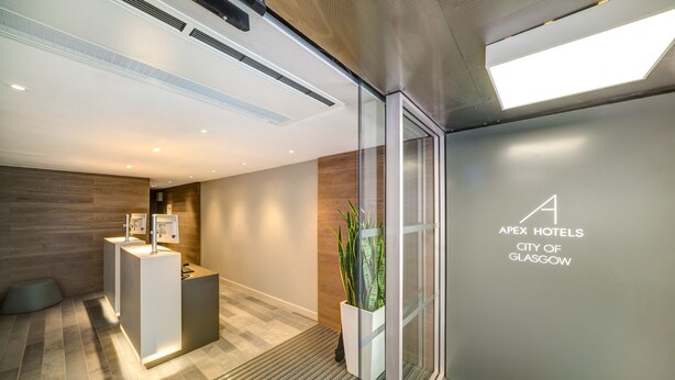 A modern hotel reception area with a reception desk, a potted plant, and a glass door leading to the lobby. - Apex City of Glasgow Hotel (Glasgow)