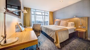 A hotel room with a large bed, a desk with a chair, a desk lamp, a bedside table with a lamp, and a view of the city through large windows. - Apex City of Glasgow Hotel (Glasgow)