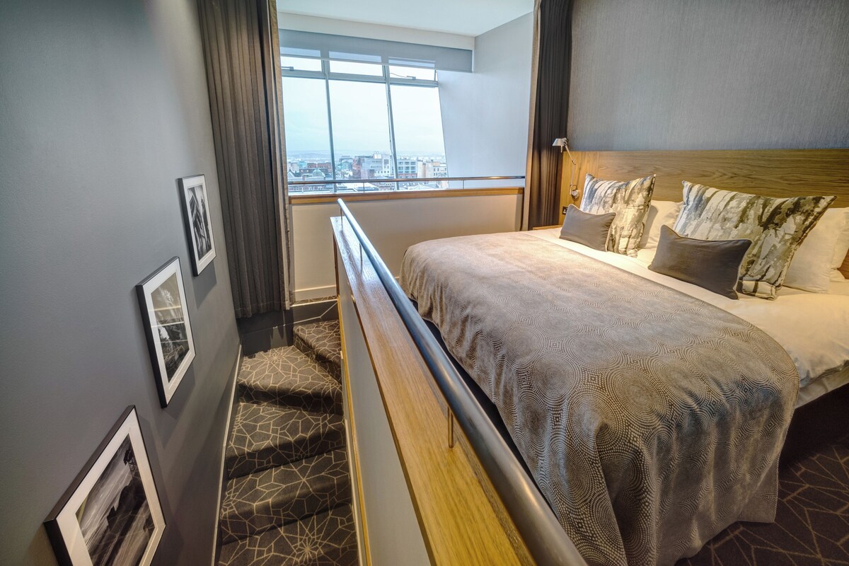 Une chambre d’hôtel avec un lit, une fenêtre donnant sur la ville, un escalier et des tableaux encadrés au mur.