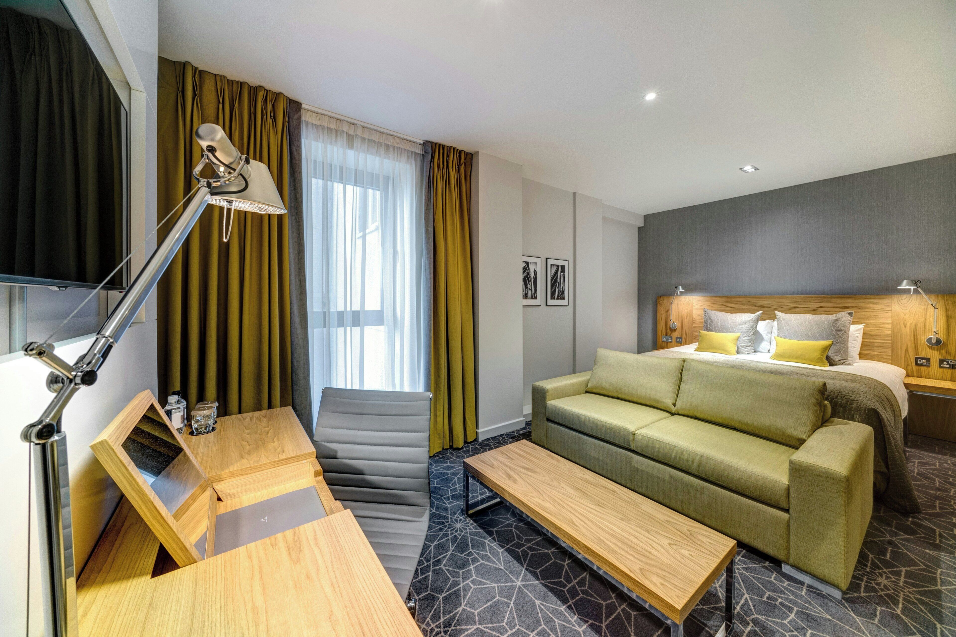 A modern hotel room with a sofa bed, a desk with a lamp, a small table, and a window with curtains.