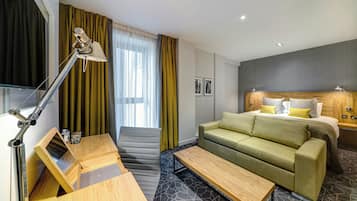 A modern hotel room with a sofa bed, a desk with a lamp, a small table, and a window with curtains.