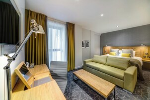 Una habitación de hotel moderna con sofá cama, escritorio con lámpara, una mesita y ventana con cortinas.