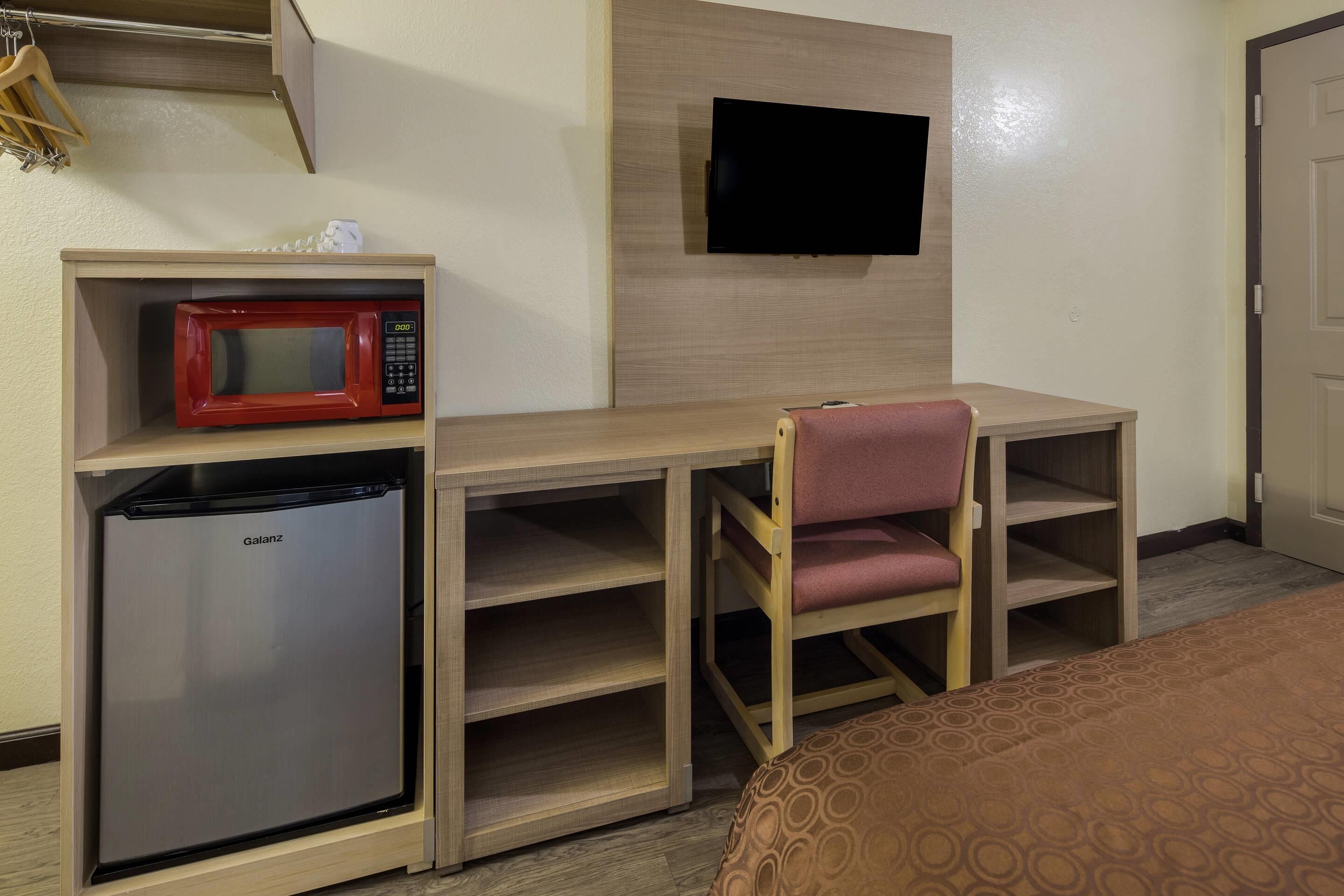 A compact hotel room with a kitchenette, a television mounted on the wall, a bed with a patterned bedspread, and a wooden desk with a chair.