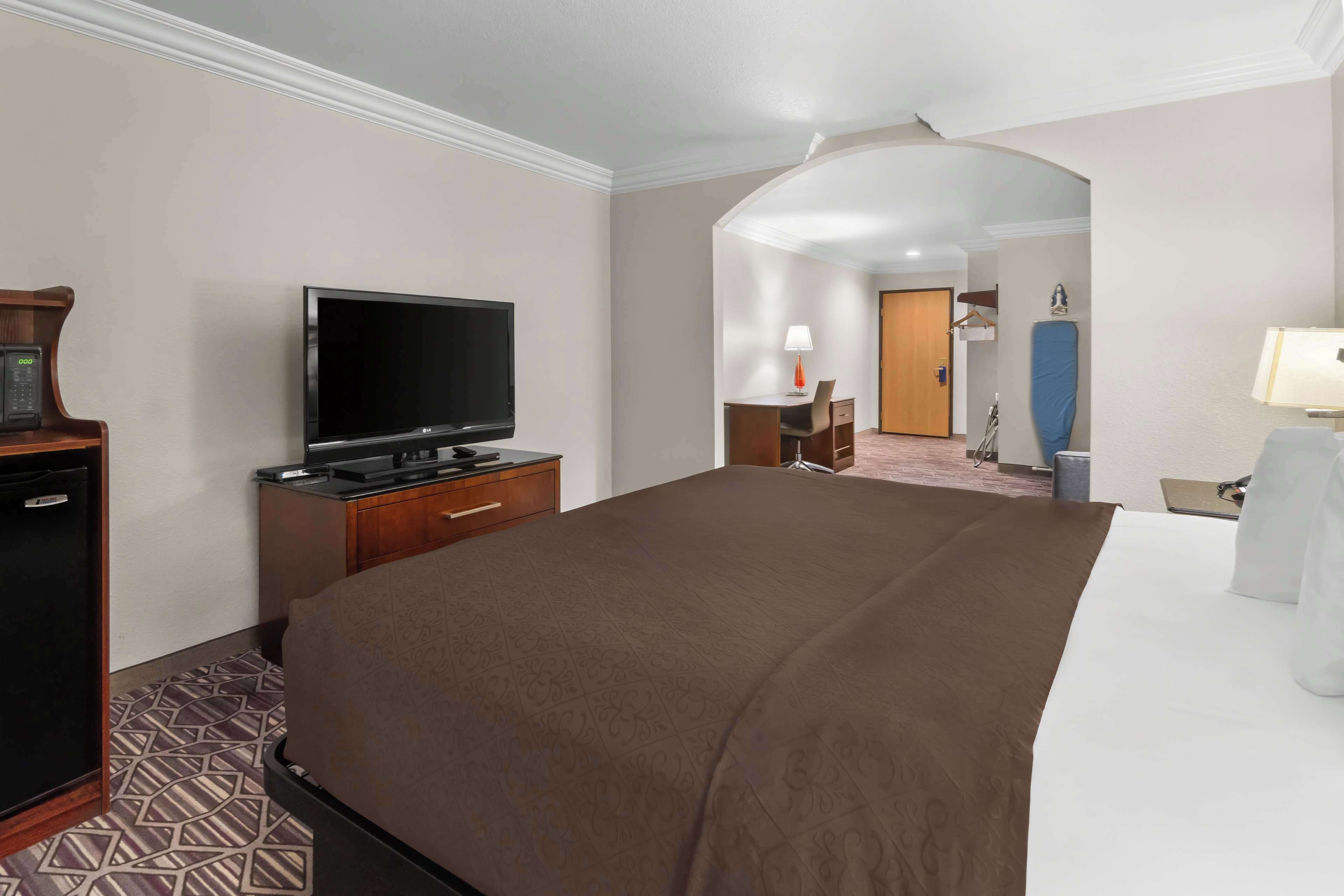 Quarto de hotel com uma cama grande, televisão de tela plana, escrivaninha com luminária e pequena geladeira.