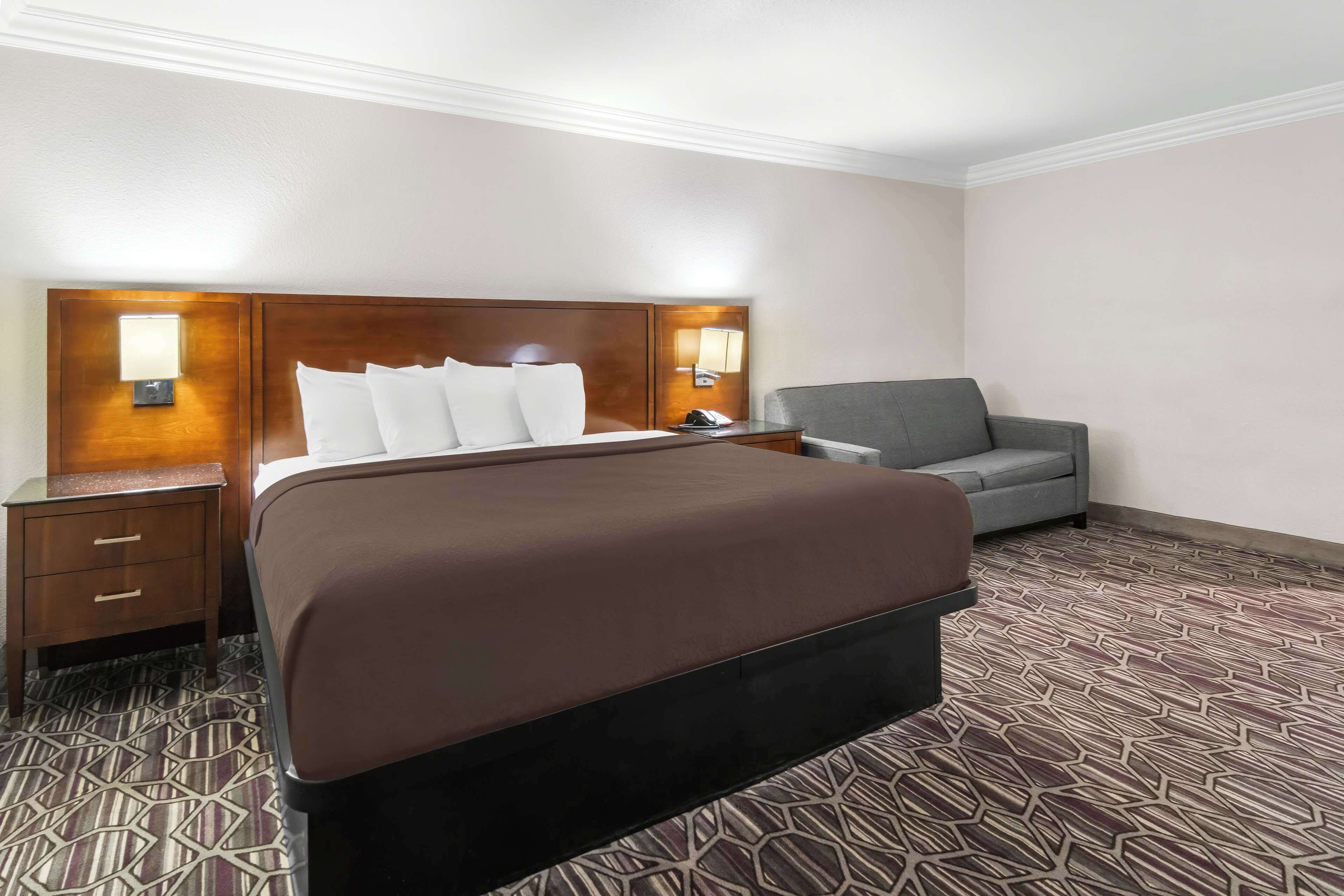 Une chambre d’hôtel avec un grand lit, un fauteuil gris, une table de chevet avec une lampe et une moquette à motifs.