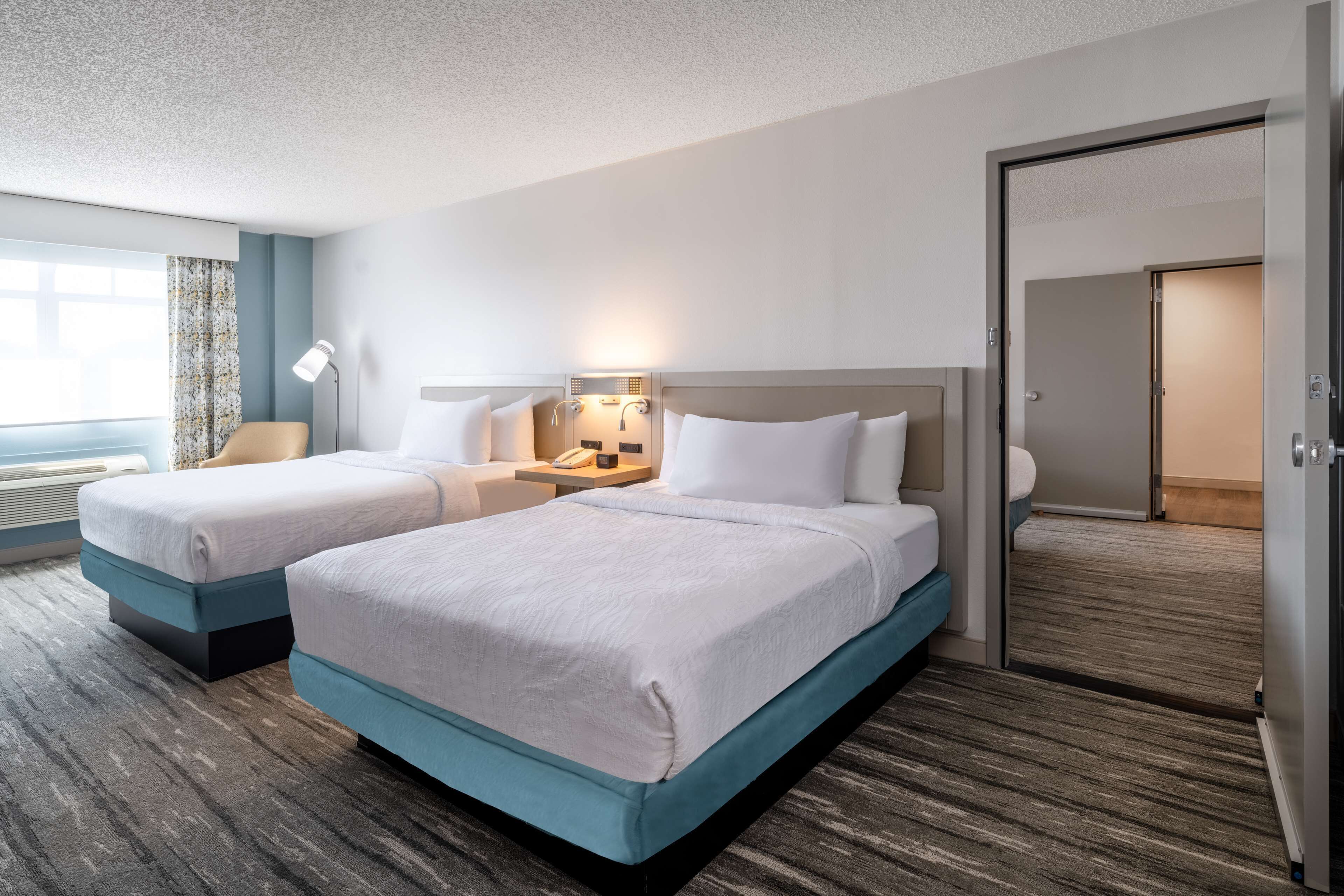 A hotel room with two beds, a desk, a lamp, and a window with curtains.