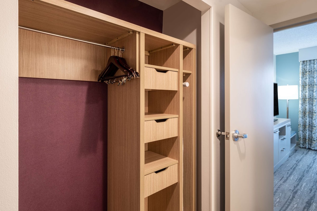 A wooden wardrobe with multiple shelves and hanging space, a door with a keyhole, and a view of a room with a bed and curtains.