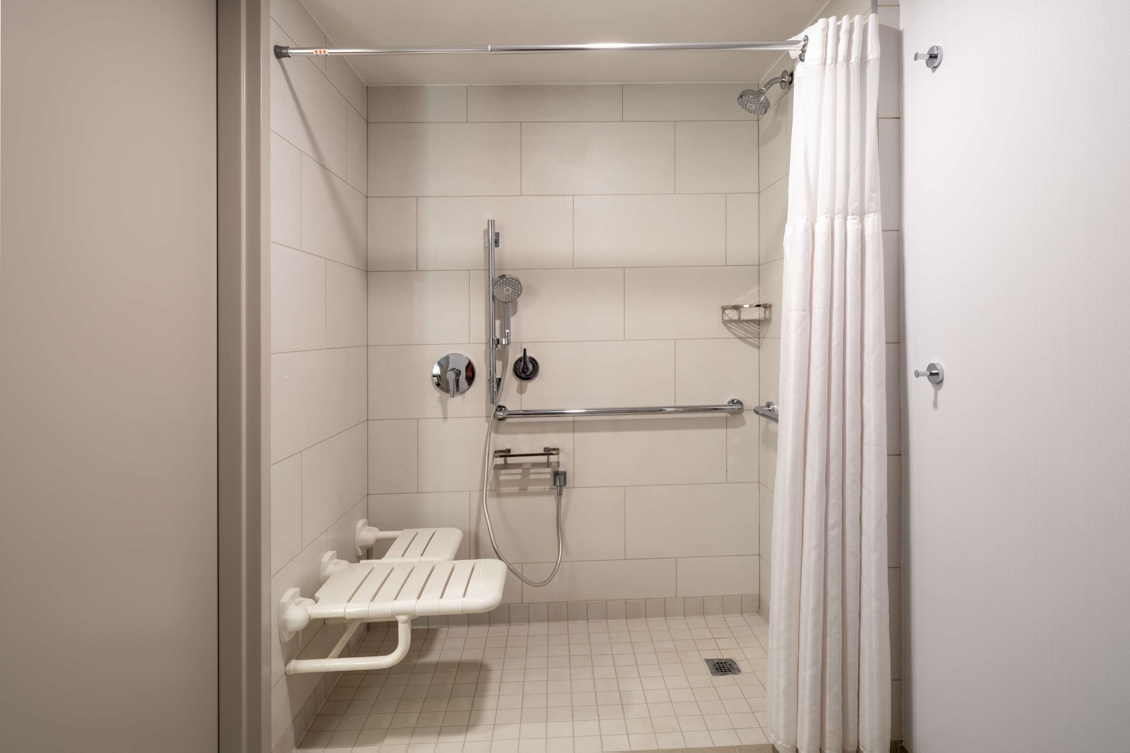A bathroom with white tiles, a shower with a handheld showerhead, a grab bar, and a white bench.