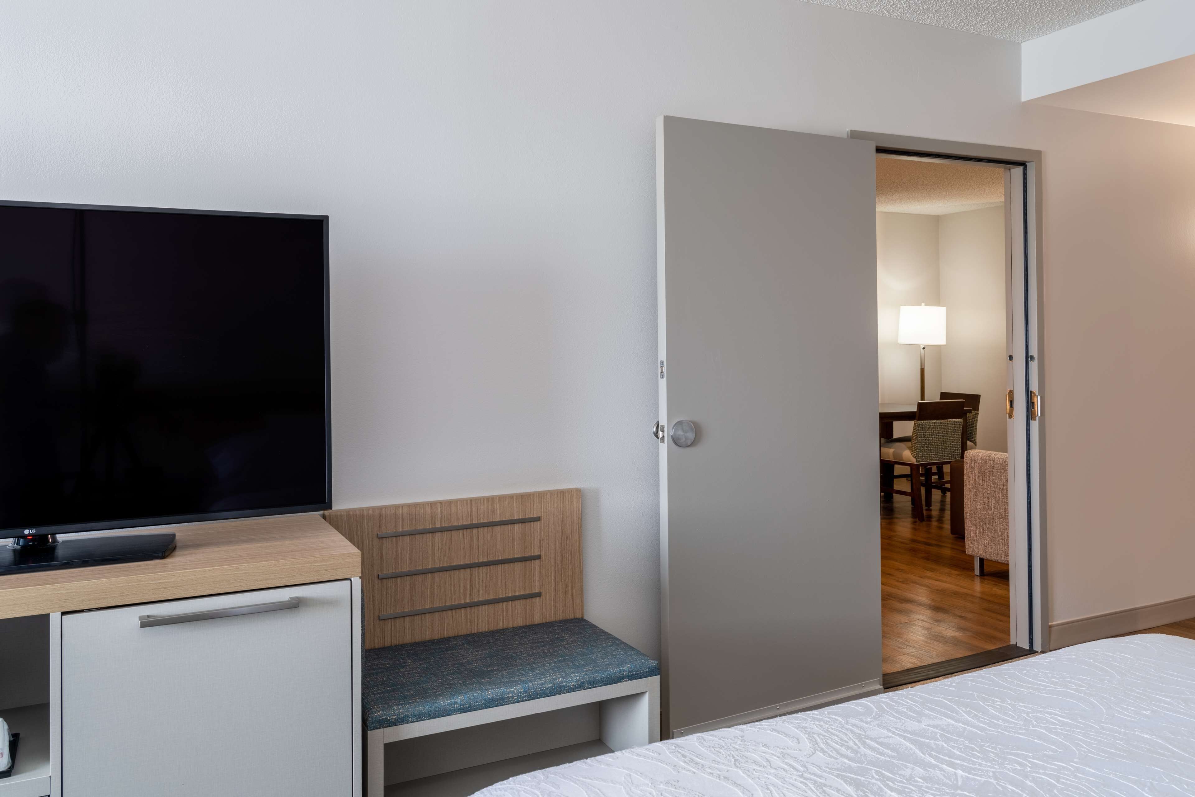 A hotel room with a bed, a TV on a desk, a wooden bench, and a door leading to a dining area.