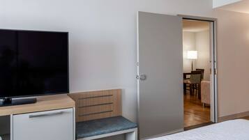 A hotel room with a bed, a TV on a desk, a wooden bench, and a door leading to a dining area.