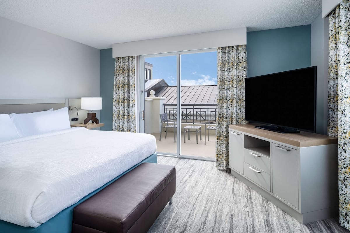 A hotel room with a large bed, a flat-screen TV, a desk, and a balcony with a table and chairs.