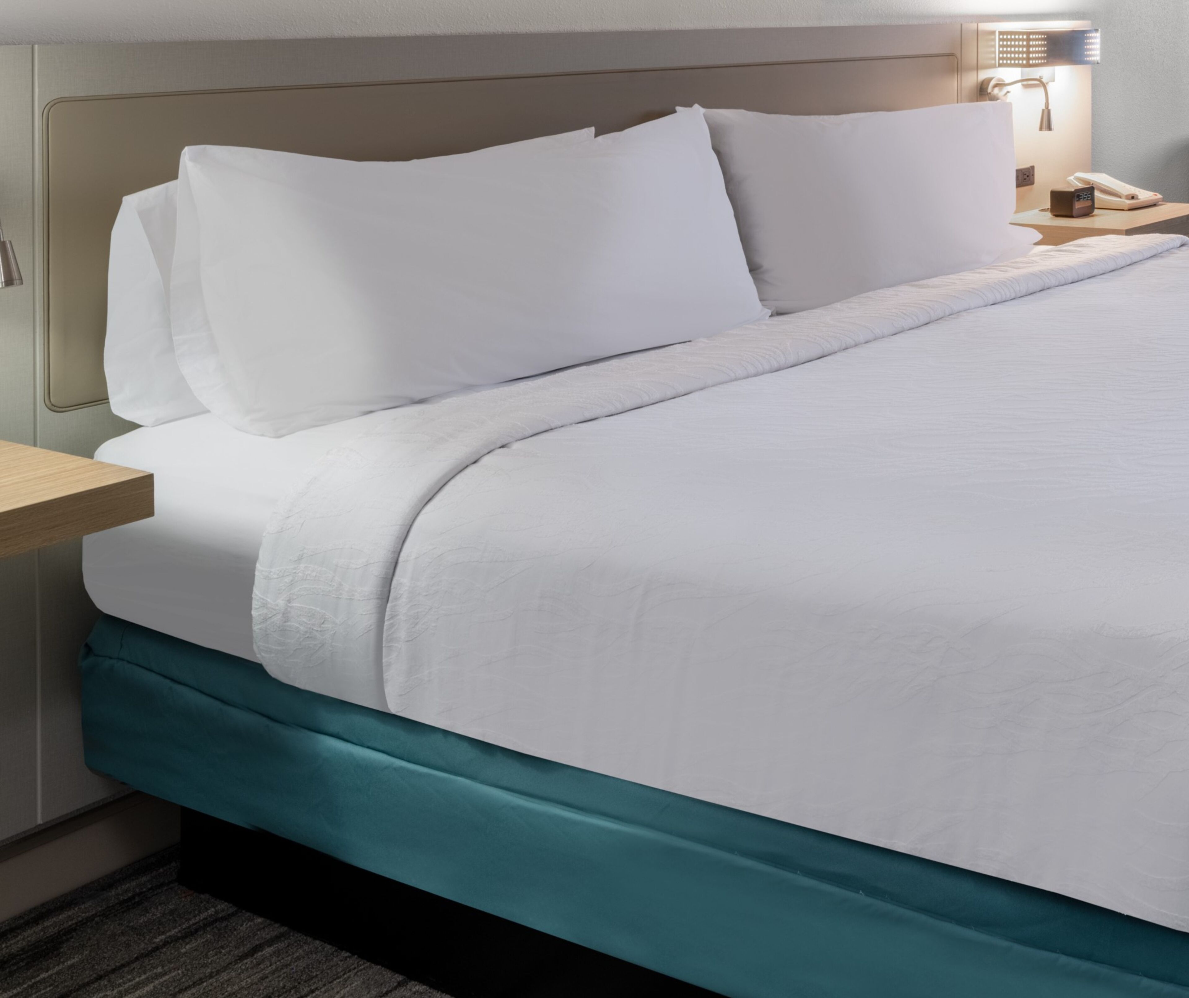 Een keurig opgemaakt bed met witte beddengoed, een blauwgroene accentkleur aan het voeteneinde, en een nachtkastje met een lamp en telefoon.
