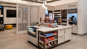 A modern coffee shop interior with a central counter, shelves stocked with snacks, and a refrigerated section labeled 'FROZEN' and 'CHILLED'. - Hilton Garden Inn Jacksonville Ponte Vedra Sawgrass (Ponte Vedra Beach)