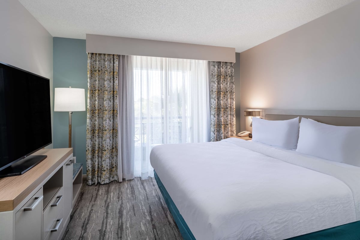 A hotel room with a large bed, a flat-screen TV, a desk with a lamp, and a view of the city through sheer curtains.