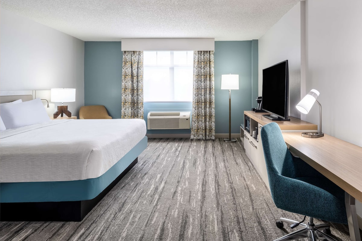 A hotel room with a bed, a desk with a chair, a television, an air conditioner, and a lamp.