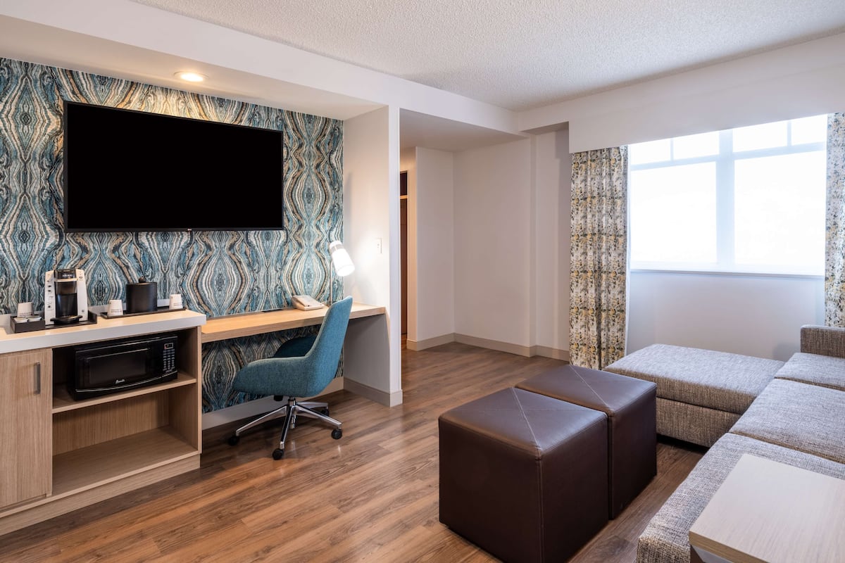A modern hotel room with a flat-screen TV, a desk with a chair, a microwave, a coffee maker, a brown ottoman, and a sofa.