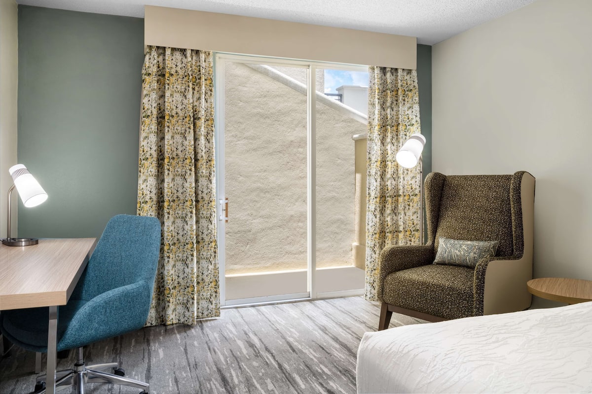 A hotel room with a desk, a blue office chair, a patterned armchair, a bed, and a sliding glass door to the outside.