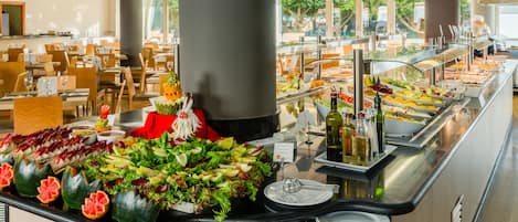 A buffet area with various food options, including salads and fruits, in a spacious dining hall with tables and chairs.