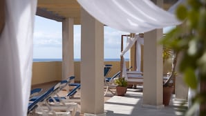 Zona de estar al aire libre iluminada por el sol, con toldos blancos y sillones reclinables azules, con vista a un cuerpo de agua.