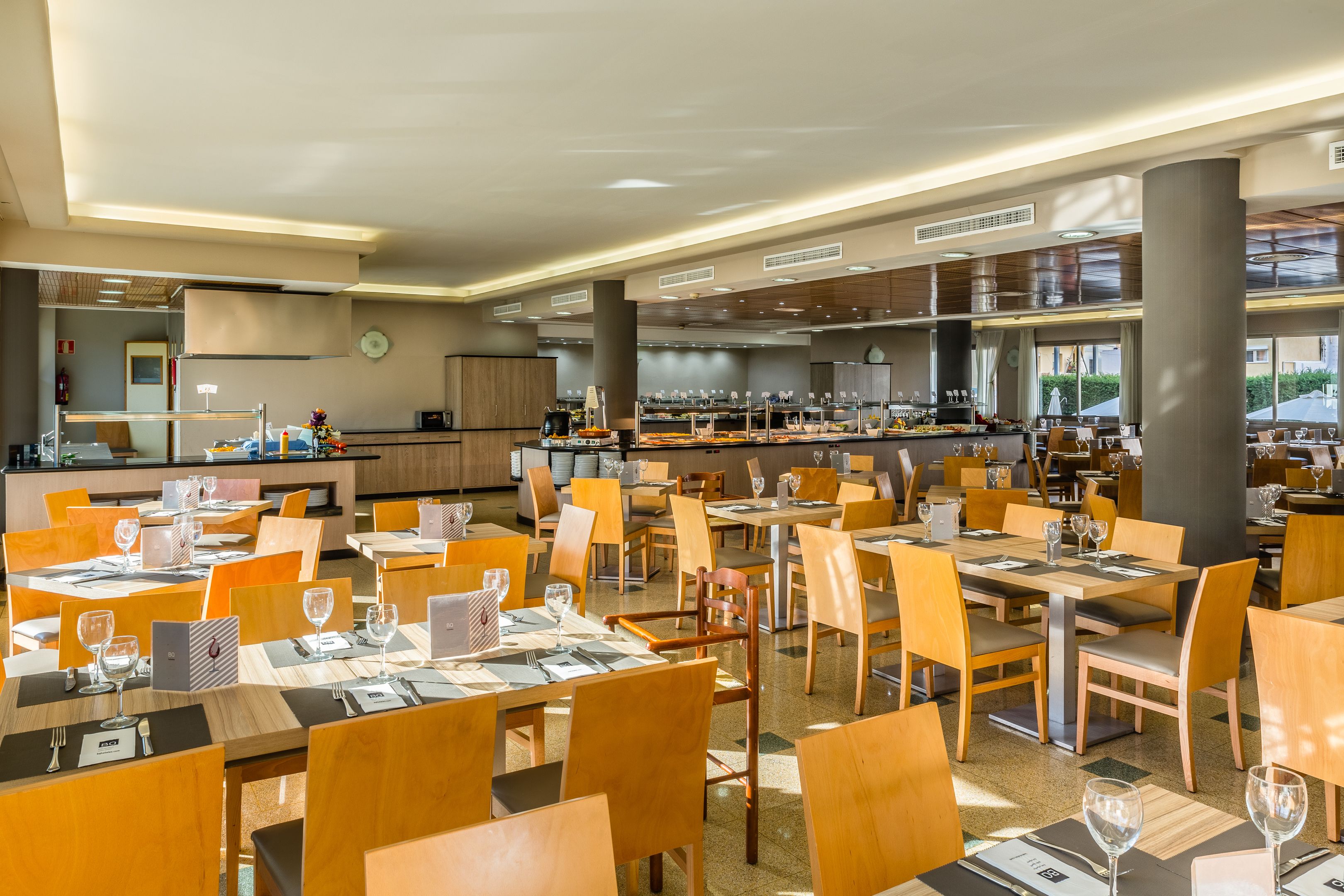 A spacious restaurant with multiple dining tables and chairs, a kitchen area, and large windows providing natural light.