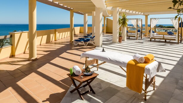 Un área de spa al aire libre con luz solar, una camilla de masaje, toallas y vistas al mar.